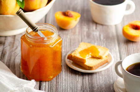 Homemade peach jam in a jar with some fresh peaches, two cups of coffee and some toasts, on a wooden table background.の写真素材