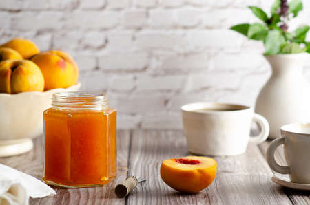 Homemade peach jam in a jar with some fresh peaches, two cups of coffee and a vase with green leaves, on a wooden table background.の写真素材