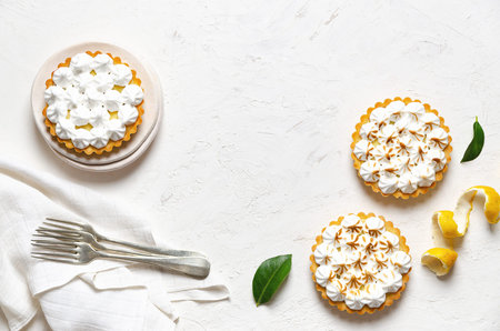 Mini lemon pies with meringue, lemon peel, lemon leaves, forks, and a white napkin, on a white background with copy space.の写真素材