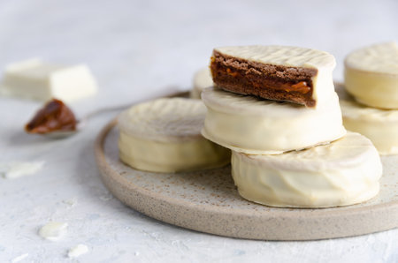 White chocolate alfajores on a beige plate, a piece of chocolate and a spoonful of dulce de leche, on light background.の写真素材