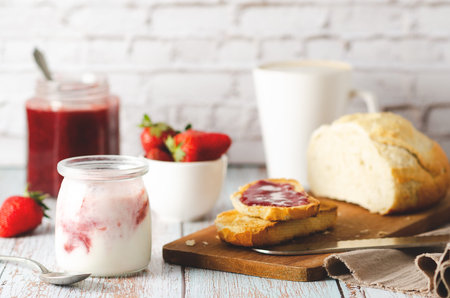 A glass of yoghurt, fresh strawberries, strawberry jam in a jar and on a slice of toasted bread, and...