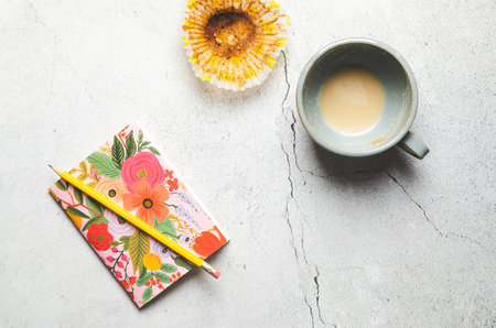 A flowered notebook with a yellow pencil, an empty cup of latte and a baking cup, on gray stoned...