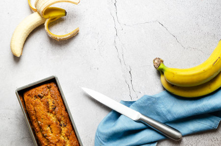 A banana bread with chocolate chips, some bananas and a blue napkin, on gray background.の写真素材