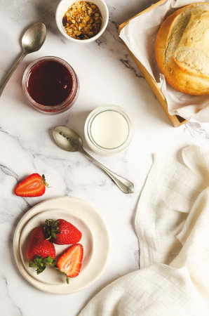 Plain yoghurt, fresh fruits, strawberry jam, a bowl of granola and a loaf of bread with a white napkin on marbled background. Topview.の写真素材