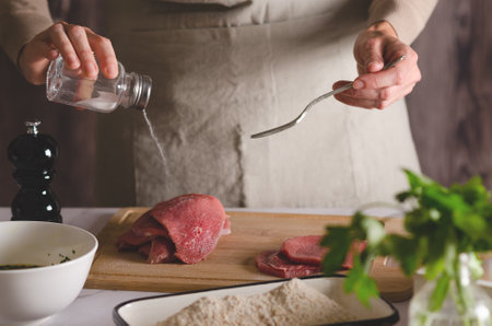 Woman in a beige apron puts salt on the meat. The process of preparing Argentine milanesas.の写真素材