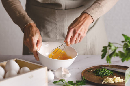 Close up of female hands whisking eggs in a bowl. Parsley and garlic on a wooden board. The process of preparing Argentine milanesas.の写真素材