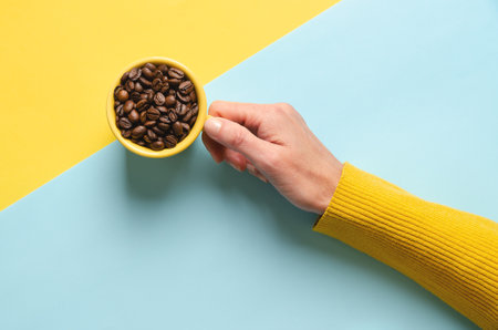 Female hand holding cup of coffee beans on blue and yellow background.の写真素材