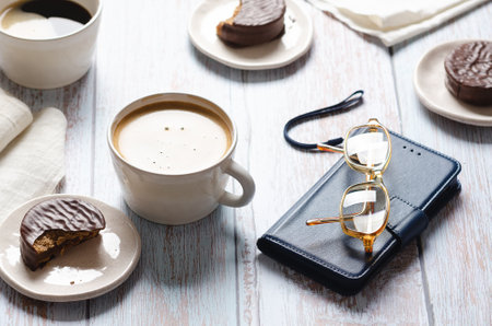 Cups of coffee, chocolate alfajores, glasses and a notebook on wooden table.の写真素材