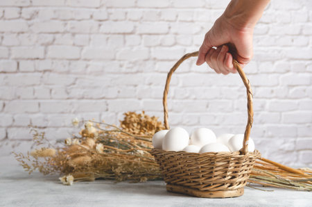 Female hand holding a basket with white chicken eggs on a light background with dried flowers. Front view with copy space.の写真素材