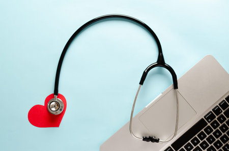 A stethoscope, a symbolic paper red heart and a laptop on a light blue background. Top view of...