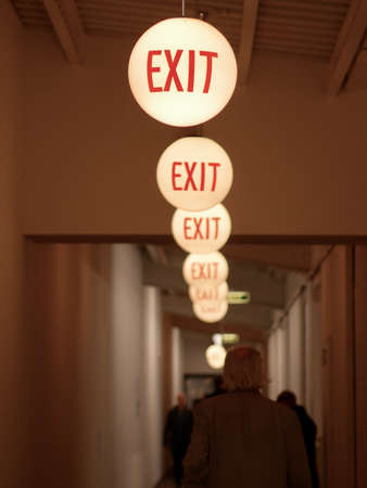 Row of round illuminated ceiling lights in a corridor or passage with walking people marking the route to the exit with red text as a safety measure in case of emergenciesの写真素材