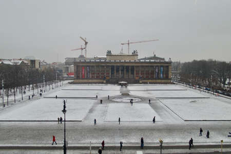 Snow over square in European city with people strolling around and tower cranes in distance, behind building with columns, winter moody day cityscapeの写真素材