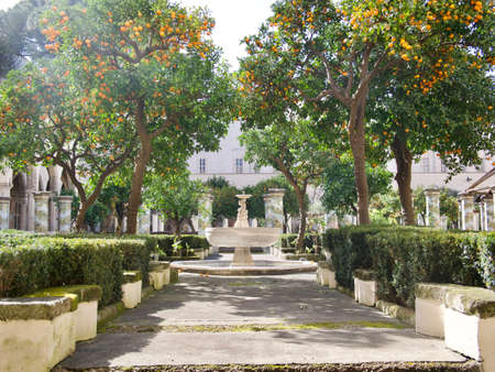 Pathway through a formal garden with fountain water feature under trees and a background of a classical historic building with colonnade of columns in frontの写真素材