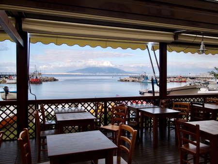 Vacant tables and chairs on a covered open-air deck at a restaurant overlooking a sheltered harbor or port with moored boatsの写真素材