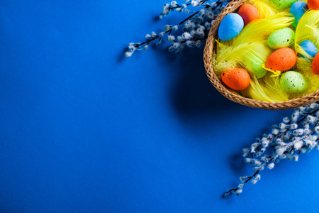 Easter Basket with easter eggs and yellow feathers and catkins on blue paper background.