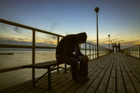 The sad guy in hood, sitting on bench at wooden pier. Baltic sea in the evening.の写真素材