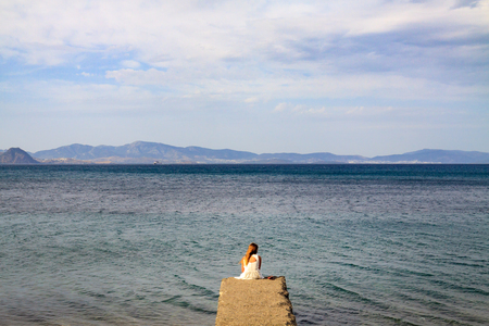 Alone woman in white casual outfit with long hair sitting at beachfront on a small concrete pier.の写真素材