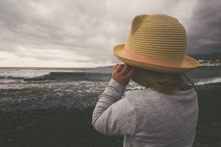 Cute little girl stays on coast of the sea in hat during stormy clouds. Vintage style photography.の写真素材