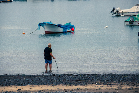 Old man with walking stick, soak his feet on coast of sea near boats in port.のeditorial素材