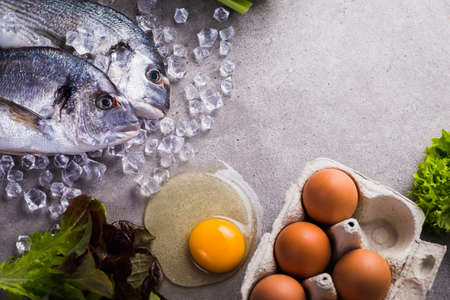 Frame of raw cooking ingredients for tasty and healthy food. Fresh dorado fish on ice, vegetables, eggs on gray concrete background top view.の写真素材
