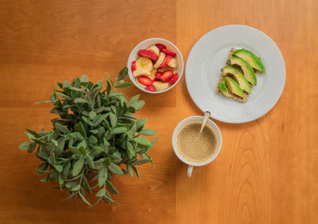 Avocado toast, cup of coffee and strawberry-banana mix in a wooden table with a decorative plantの写真素材