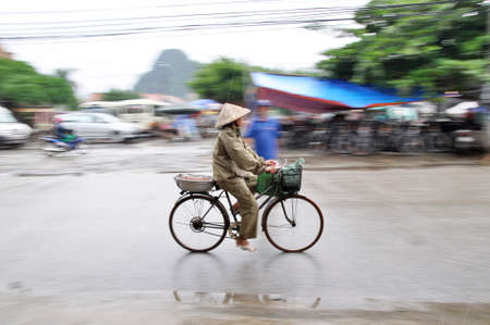 Hanoi, Vietnam - Sept 2013: A panning shot of a Hanoi lady wearing a straw hat cycling in the rain.のeditorial素材