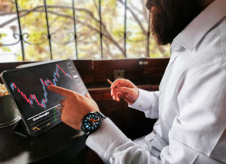 young adult bearded businessman with smart watch smoking cigar and touching his pro tablet screen checking market stock exchange for-ex block chain graphicの写真素材