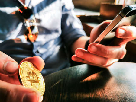 young adult male with sunglasses and watch using his smartphone and flipping gold bitcoin on the tableの写真素材