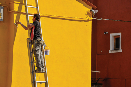 Burano, Italy - May 08, 2013. Close-up of painter painting wall of yellow house on sunny day in Burano, a gracious little town full of canals, near Venice. In the Veneto region, northern Italyのeditorial素材