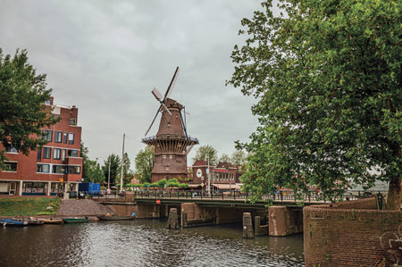 Bridge over canal, trees and old windmill on the bank with cloudy sky in Amsterdam. Famous for its...