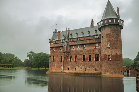De Haar Castle facade with ornate brick towers and water moat on rainy day, near Utrecht. Of...