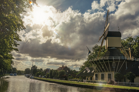 Wide tree-lined canal with windmill and boats and shine of sunset reflected in water at Weesp. Quiet and pleasant village full of canals and green near Amsterdam. Northern Netherlands. Retouched photoの写真素材