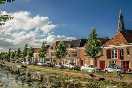 Weesp, northern Netherlands - June 26, 2017. Tree-lined canal with aquatic plants, church bell tower and brick houses at the bank on sunset in Weesp. Quiet and pleasant village full of canals and green near Amsterdam.のeditorial素材