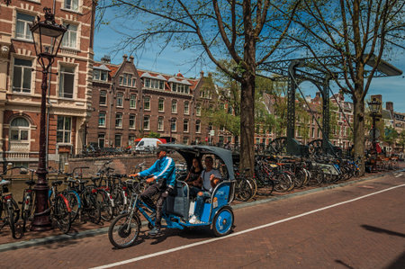 Amsterdam, northern Netherlands - June 27, 2017. Rickshaw riding on street beside canal and old...