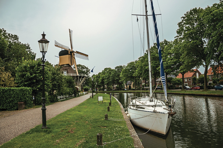 Boat and wooden yellow windmill next to wide tree-lined canal under cloudy sky at sunset in Weesp. Quiet and pleasant village full of canals and green near Amsterdam. Northern Netherlands.の写真素材