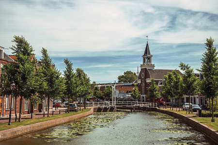 Tree-lined canal with bascule bridge, church and brick houses in street on the banks on sunny day in Weesp. Quiet and pleasant village full of canals and green near Amsterdam. Northern Netherlands.の写真素材