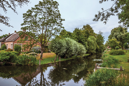 Charming small canal next to rustic house with lush garden and trees reflected on the water in cloudy day at Drimmelen. A lovely small hamlet with harbor and elegance streets. Southern Netherlands.の写真素材