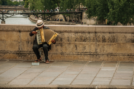 Paris, northern France - July 12, 2017. Old man with hat playing accordion over bridge at the Seine River on cloudy sky in Paris. Known as one of the world's most impressive cultural centers.のeditorial素材