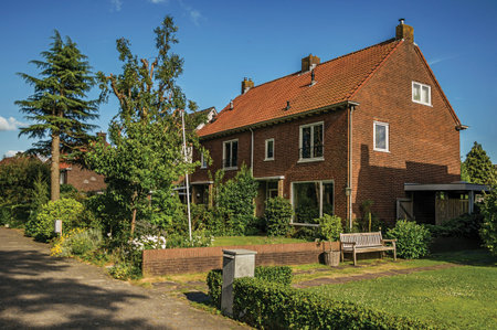 Orange brick house with beautiful and verdant garden in front of alley under blue sky at Weesp. Quiet and pleasant village full of canals and green near Amsterdam. Northern Netherlands.のeditorial素材
