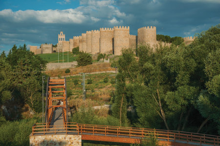Wooden bridge over the Adaja River with trees and the large wall encircling the town of Avila at...
