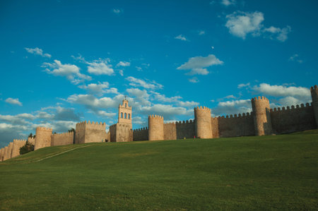 Stone large wall with towers over hilly landscape covered by lawn, encircling the Avila town at...