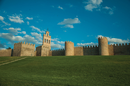 Stone large wall with towers over hilly landscape covered by lawn, encircling the Avila town at sunset. It has the longest and imposing wall completely encircling this well-kept gothic town in Spain.のeditorial素材