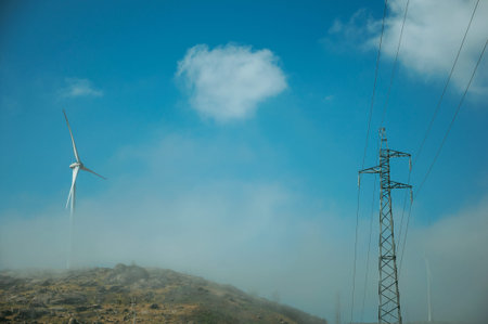 Wind turbines for electric power generation on hill with mist next to electric tower, in a sunny day...