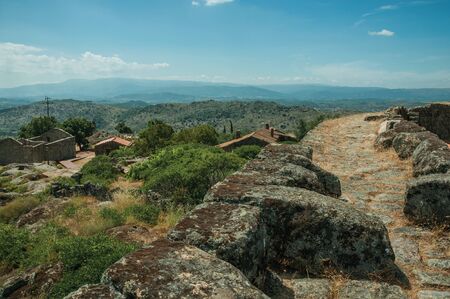 Pathway over thick stone wall next to old houses roofs and verdant hilly landscape, in a sunny day at Sortelha. One of the most astonishing and well preserved medieval villages in all Portugal.の写真素材