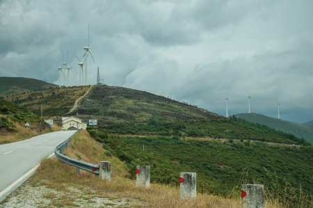 Countryside road passing through hilly landscape with several wind turbines, in a cloudy day at...