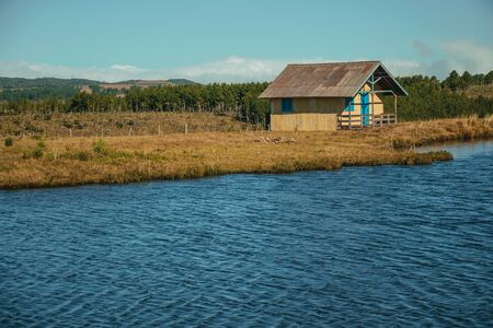 Charming wooden chalet facing a blue water lake in a hilly landscape with trees and dry bushes near Cambara do Sul. A small rural town in southern Brazil with amazing natural tourist attractions.の写真素材