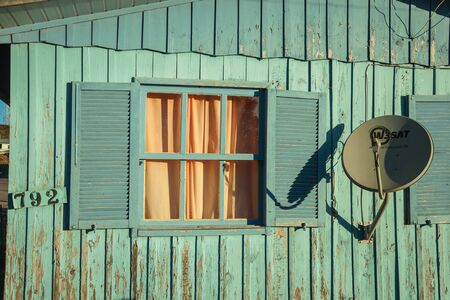 Cambara do Sul, Brazil - July 16, 2019. Modern satellite dish and open window in old worn wooden house at sunset in Cambara do Sul. A small country town with amazing natural tourist attractions.のeditorial素材