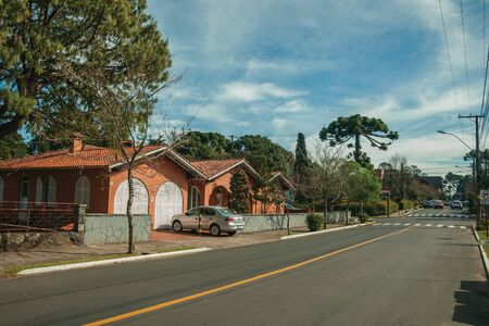 Gramado, Brazil - July 21, 2019. Gardened houses and trees on empty paved street with parked cars in a sunny day at Gramado. A cute european-influenced town highly sought after by tourists.のeditorial素材