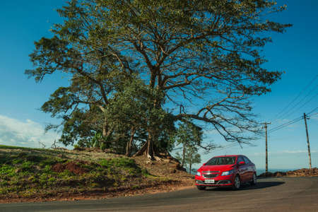 Pardinho, Brazil - June 20, 2019. Car going through paved country road beside leafy tree on a slope covered by green meadow near Pardinho. A small rural village in the countryside of Sao Paulo State.のeditorial素材