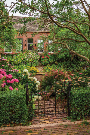 Charming rustic brick house with flowered garden, leafy trees and iron gate in a cloudy day at Drimmelen. A lovely small hamlet in Netherlands.の写真素材